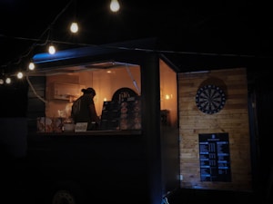 A dimly lit food truck is decorated with hanging string lights. Inside, a person works at the counter, surrounded by menus and cooking equipment. The exterior features a dartboard mounted on a wooden wall, contributing to the cozy, informal atmosphere.