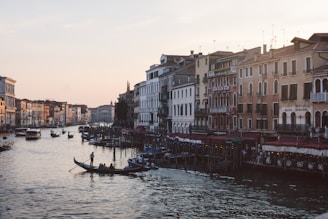 A scenic canal is flanked by historic buildings with intricate architectural details. Gondolas glide along the water, carrying passengers enjoying the view. The atmosphere suggests a serene evening setting, as the warm glow of lights begins to emanate from the waterside cafes and restaurants.