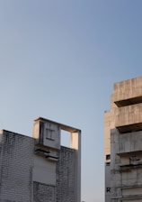 Two modern, concrete buildings with textured facades, featuring minimal architectural details and clean lines. The right building has a partially visible letter 'R' and oversized balconies. The sky is clear with a soft, warm light illuminating the structures.