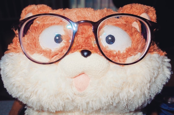 A plush toy resembling an animal with a fluffy texture, wearing oversized eyeglasses. The toy features a mix of brown and cream colors, and has a playful, surprised expression with big eyes.