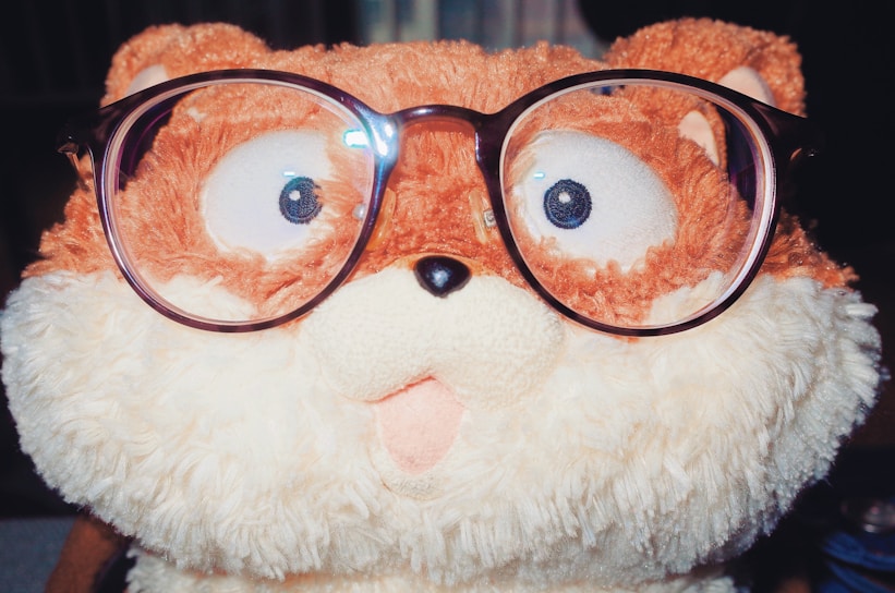 A plush toy resembling an animal with a fluffy texture, wearing oversized eyeglasses. The toy features a mix of brown and cream colors, and has a playful, surprised expression with big eyes.