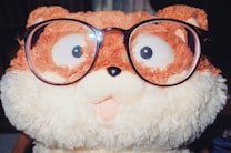 A plush toy resembling an animal with a fluffy texture, wearing oversized eyeglasses. The toy features a mix of brown and cream colors, and has a playful, surprised expression with big eyes.