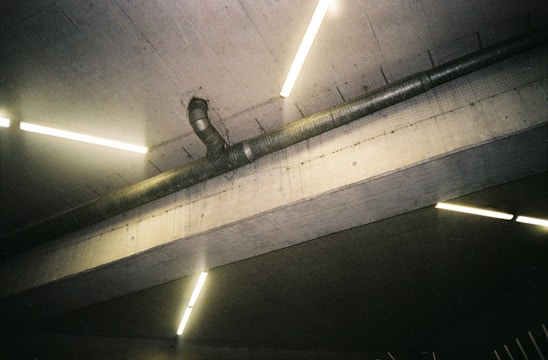 A concrete ceiling with exposed ventilation pipes is intersected by bright, fluorescent tube lights. The image captures an industrial aesthetic with its raw, unfinished surfaces.