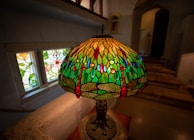 A Tiffany-style stained glass lamp with a colorful dragonfly motif is prominently displayed on a table. The lampshade features intricate designs with shades of green, red, blue, and yellow, creating a vibrant and artistic appearance. In the background, a window with stained glass patterns and a stone staircase are visible, contributing to an antique and cozy ambiance.