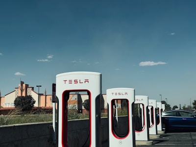 A row of Tesla charging stations is lined up in a parking area, with a modern building in the background. A Tesla car is positioned next to one of the chargers, ready for charging. The sky is predominantly clear, with a few small clouds adding texture.
