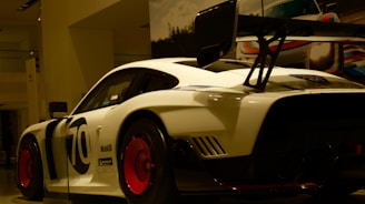 A sleek white race car with bold black lettering and a large rear spoiler sits on display, illuminated under showroom lights. The vehicle features prominent red wheels and various sponsor logos.