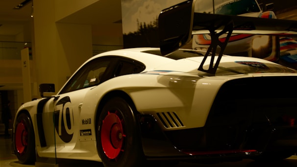 A sleek white race car with bold black lettering and a large rear spoiler sits on display, illuminated under showroom lights. The vehicle features prominent red wheels and various sponsor logos.