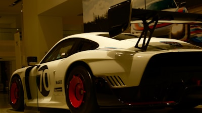 A sleek white race car with bold black lettering and a large rear spoiler sits on display, illuminated under showroom lights. The vehicle features prominent red wheels and various sponsor logos.