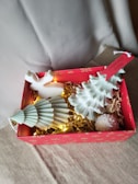 A festive gift box with a red exterior decorated with gold snowflakes contains decorative items. Inside are two white, Christmas tree-shaped candles, a small reindeer ornament, straw filler, and warm white string lights that add a cozy glow. A hanging tag with 'Merry Christmas' adds to the festive theme.