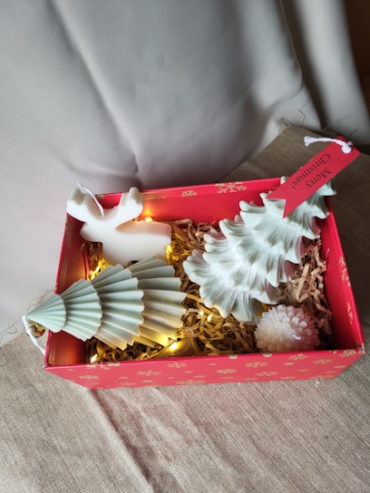 A festive gift box with a red exterior decorated with gold snowflakes contains decorative items. Inside are two white, Christmas tree-shaped candles, a small reindeer ornament, straw filler, and warm white string lights that add a cozy glow. A hanging tag with 'Merry Christmas' adds to the festive theme.