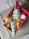 A decorative Christmas-themed box containing artistic candles shaped like trees and a bird, placed on straw-like material. The candles are white and green, accompanied by warm yellow fairy lights. A red tag with 'Merry Christmas!' hangs from one of the tree candles.
