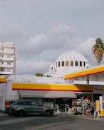 A gas station with a modern design featuring a prominent red and yellow color scheme. In the foreground, there is a dark-colored SUV parked next to a small convenience store displaying various products. The background shows a white-domed building, likely a church, and some palm trees, adding a contrast between urban and traditional architectural elements.