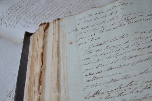 An aged, open book with handwritten text in flowing script lies on a surface. The pages are yellowed and worn, suggesting antiquity and frequent handling. The handwriting is elegant and cursive, adding a historic and personal touch.