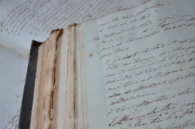 An aged, open book with handwritten text in flowing script lies on a surface. The pages are yellowed and worn, suggesting antiquity and frequent handling. The handwriting is elegant and cursive, adding a historic and personal touch.