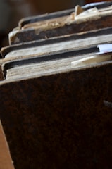 A close-up view of a stack of old, weathered books with hard, textured covers. The spines show signs of wear and age, with frayed edges and discolored pages. Several paper bookmarks protrude from between the pages, indicating use or saved spots.