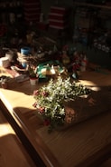 A wooden table is adorned with various crafting supplies such as ribbons, small potted plants, cups, and decorative items. In the foreground, a bouquet wrapped in newspaper features greenery and vibrant red berries. The soft sunlight casts shadows and highlights the natural textures of the wood and plants, creating a cozy and warm atmosphere.