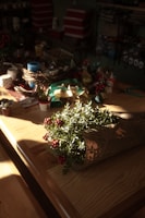 A wooden table is adorned with various crafting supplies such as ribbons, small potted plants, cups, and decorative items. In the foreground, a bouquet wrapped in newspaper features greenery and vibrant red berries. The soft sunlight casts shadows and highlights the natural textures of the wood and plants, creating a cozy and warm atmosphere.