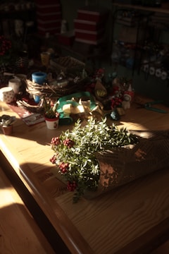 A wooden table is adorned with various crafting supplies such as ribbons, small potted plants, cups, and decorative items. In the foreground, a bouquet wrapped in newspaper features greenery and vibrant red berries. The soft sunlight casts shadows and highlights the natural textures of the wood and plants, creating a cozy and warm atmosphere.