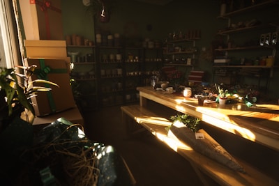 A cozy room filled with natural light features several shelves stocked with various flower pots and gardening supplies. A large wooden table occupies the center, scattered with small potted plants, wrapping paper, and decorative items. On the left, several stacked gift boxes are adorned with ribbons, and in the background, more pots and planters can be seen.