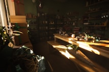 A cozy room filled with natural light features several shelves stocked with various flower pots and gardening supplies. A large wooden table occupies the center, scattered with small potted plants, wrapping paper, and decorative items. On the left, several stacked gift boxes are adorned with ribbons, and in the background, more pots and planters can be seen.