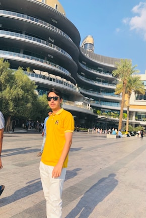 A person wearing sunglasses and a bright yellow polo shirt stands in an outdoor area with a modern, multi-level building in the background. The building features several restaurant names and has a sleek, curved design. There are palm trees and people walking around, with a clear blue sky overhead.