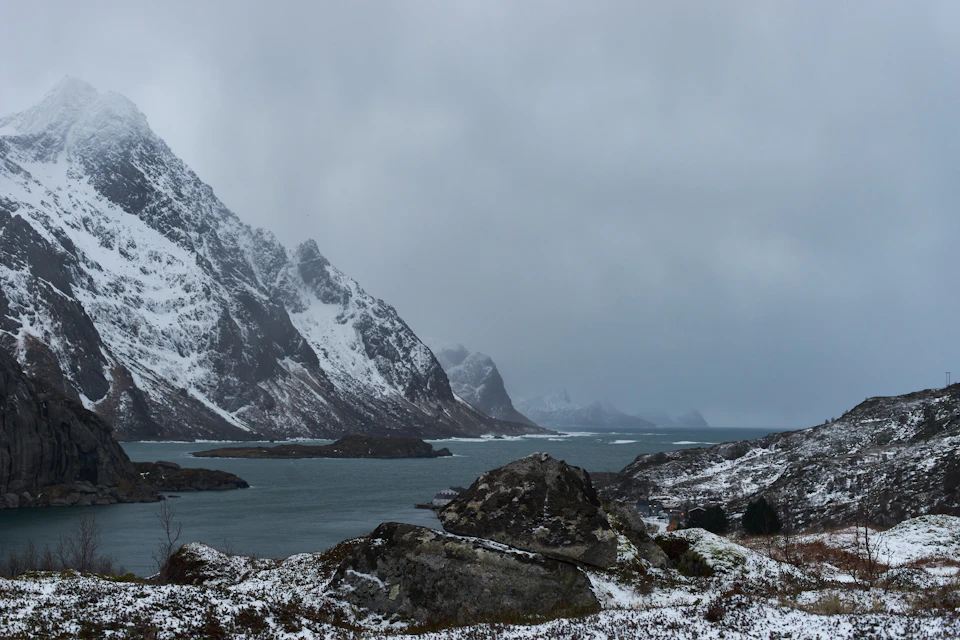 The image depicts a serene winter landscape with snow-covered mountains and a large body of water, possibly an ocean or lake, nestled between them. The sky above is overcast, casting a soft light on the scene below. In the foreground, there are rocks scattered across the ground, adding texture to the otherwise smooth terrain. The predominant colors in the image are shades of gray from the snow and the overcast sky, with hints of blue and green from the water and vegetation.