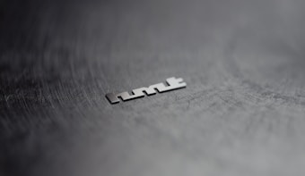A metallic object is placed on a dark, textured surface. The object appears to be a word or logo consisting of distinct, angular letters. The background is blurred, enhancing the focus on the object.