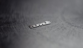 A metallic object is placed on a dark, textured surface. The object appears to be a word or logo consisting of distinct, angular letters. The background is blurred, enhancing the focus on the object.