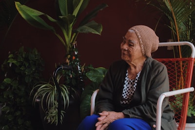 An elderly woman wearing a knitted hat and a dark green cardigan is sitting on a red and white chair in a room adorned with various potted plants, including tall leafy plants and vines.