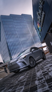 A sleek silver Lexus SUV is parked in front of a modern glass skyscraper on a wet, patterned brick pavement. The overcast sky creates a moody atmosphere complemented by reflections on the building's windows. The logo of a company is visible on the right, adding a touch of urban professionalism.