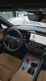 The interior of a luxury car showcasing a sleek and modern design. A leather steering wheel with a prominent logo, a large touchscreen display with various controls, and elegant leather seats are visible. The dashboard has a sophisticated and stylish appearance with high-quality materials. Outside, a snowy path and part of a brick building can be seen through the windshield.
