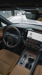 The interior of a luxury car showcasing a sleek and modern design. A leather steering wheel with a prominent logo, a large touchscreen display with various controls, and elegant leather seats are visible. The dashboard has a sophisticated and stylish appearance with high-quality materials. Outside, a snowy path and part of a brick building can be seen through the windshield.