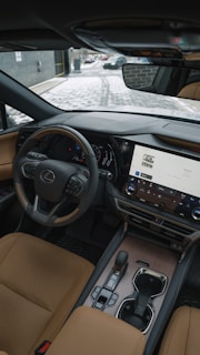 The interior of a luxury car showcasing a sleek and modern design. A leather steering wheel with a prominent logo, a large touchscreen display with various controls, and elegant leather seats are visible. The dashboard has a sophisticated and stylish appearance with high-quality materials. Outside, a snowy path and part of a brick building can be seen through the windshield.