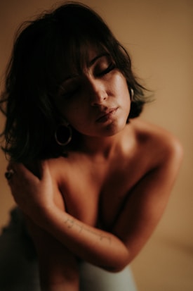 A woman is captured in a close-up, intimate pose with her eyes closed. She appears to be nude or wearing very little, with her arms wrapping around herself in a protective manner. The background is neutral, and the lighting is soft, focusing on her face and upper body.