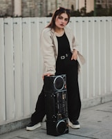 A woman stands confidently with one hand resting on a large boombox. She wears a stylish beige jacket over a black outfit, complete with a belt and matching black shoes. Sunglasses are perched on her head, and her expression is poised and self-assured. The backdrop features a white fence and an urban landscape, creating a modern city vibe.
