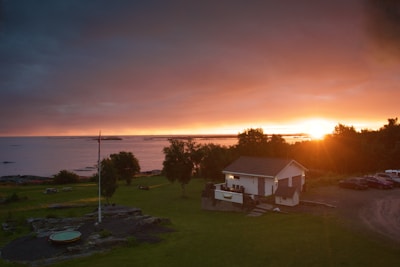 A serene landscape featuring a house and several cars parked on a grassy area near a body of water. The sun is setting in the background, casting an orange glow across the scene. Trees and the shoreline are visible in the distance. A flagpole and a circular stone structure are also present.