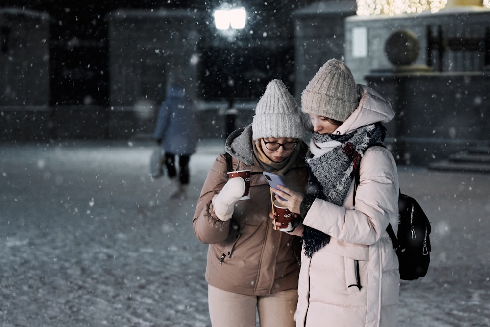 two women standing in the snow looking at a cell phone