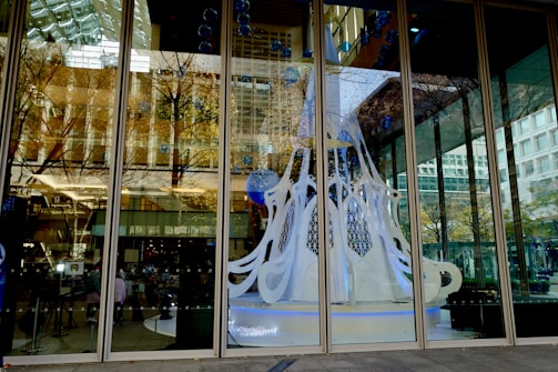 A modern architectural space with large glass windows reflecting urban buildings and trees. Inside, a white and blue artistic structure resembling a stylized Christmas tree is displayed, accentuated by lights and hanging decorations.