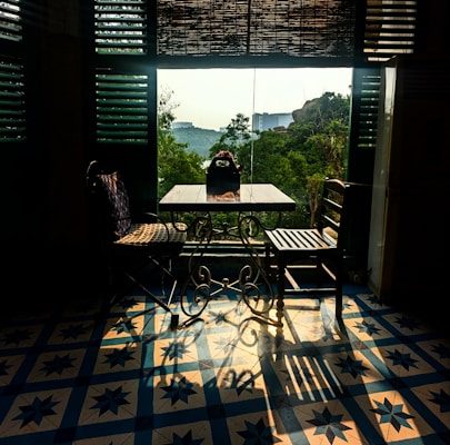 A cozy seating area with a small table and two chairs, one wicker and one wooden, bathed in warm sunlight filtering through wooden blinds. The floor features a patterned tile design in blue and white. Outside the window, lush green trees and a distant view of modern buildings are visible under soft daylight.