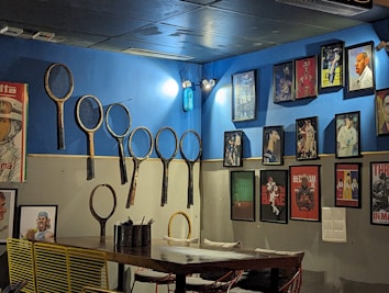 A room with blue and white walls adorned with a collection of vintage tennis rackets and framed sports photographs. The rackets are mounted on the wall, while various sports-themed artworks display athletes in action. A wooden table with yellow wire chairs and some utensils sits beneath the display.