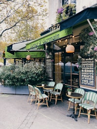 A cozy outdoor café with green awnings and bistro-style tables and chairs. The entrance is surrounded by potted plants and decorative elements, creating a welcoming and charming atmosphere. The menu board on the wall lists various drinks available.
