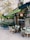 A cozy outdoor café with green awnings and bistro-style tables and chairs. The entrance is surrounded by potted plants and decorative elements, creating a welcoming and charming atmosphere. The menu board on the wall lists various drinks available.