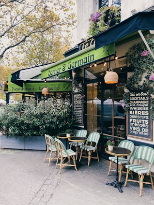 A cozy outdoor café with green awnings and bistro-style tables and chairs. The entrance is surrounded by potted plants and decorative elements, creating a welcoming and charming atmosphere. The menu board on the wall lists various drinks available.