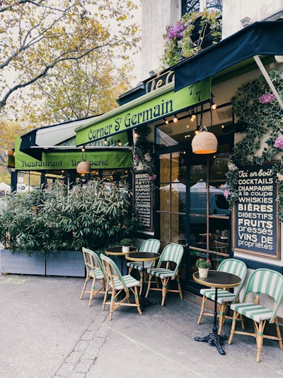 A cozy outdoor café with green awnings and bistro-style tables and chairs. The entrance is surrounded by potted plants and decorative elements, creating a welcoming and charming atmosphere. The menu board on the wall lists various drinks available.
