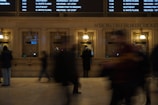A busy indoor scene at a train station ticket counter with people in motion, indicating activity and movement. The ticket booths are designed with classic architecture, featuring ornate details and warm lighting. There is a large digital departure board displaying train times above the counters.