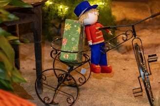 A decorative doll dressed in a red and blue outfit with gold accents is seated on a vintage mini bicycle made of wrought iron. Next to the doll is a green gift wrapped in festive paper with star patterns. The background features warm lighting and greenery, creating a cozy ambiance.