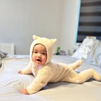 A baby wearing a fluffy beige onesie with small ears is lying on a bed. The room is softly lit, with a window featuring black-and-white striped blinds in the background. The bedding is light-colored, and there are some items visible on a surface in the corner of the room.