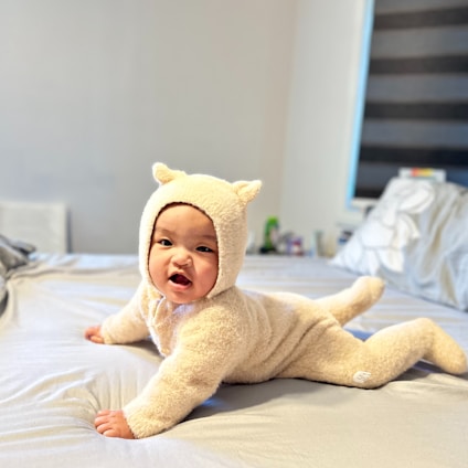 A baby wearing a fluffy beige onesie with small ears is lying on a bed. The room is softly lit, with a window featuring black-and-white striped blinds in the background. The bedding is light-colored, and there are some items visible on a surface in the corner of the room.