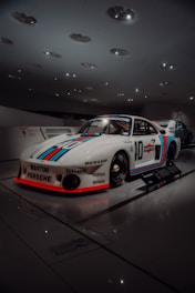 A sleek racing car displayed in a well-lit showroom, featuring distinctive racing stripes in red and blue over a white body. The car is branded with several logos including Martini, Porsche, and others, with a classic and aerodynamic design. The surrounding environment has a modern and minimalist architecture with ceiling lights creating a sophisticated ambiance.