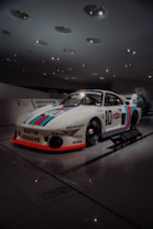 A sleek racing car displayed in a well-lit showroom, featuring distinctive racing stripes in red and blue over a white body. The car is branded with several logos including Martini, Porsche, and others, with a classic and aerodynamic design. The surrounding environment has a modern and minimalist architecture with ceiling lights creating a sophisticated ambiance.